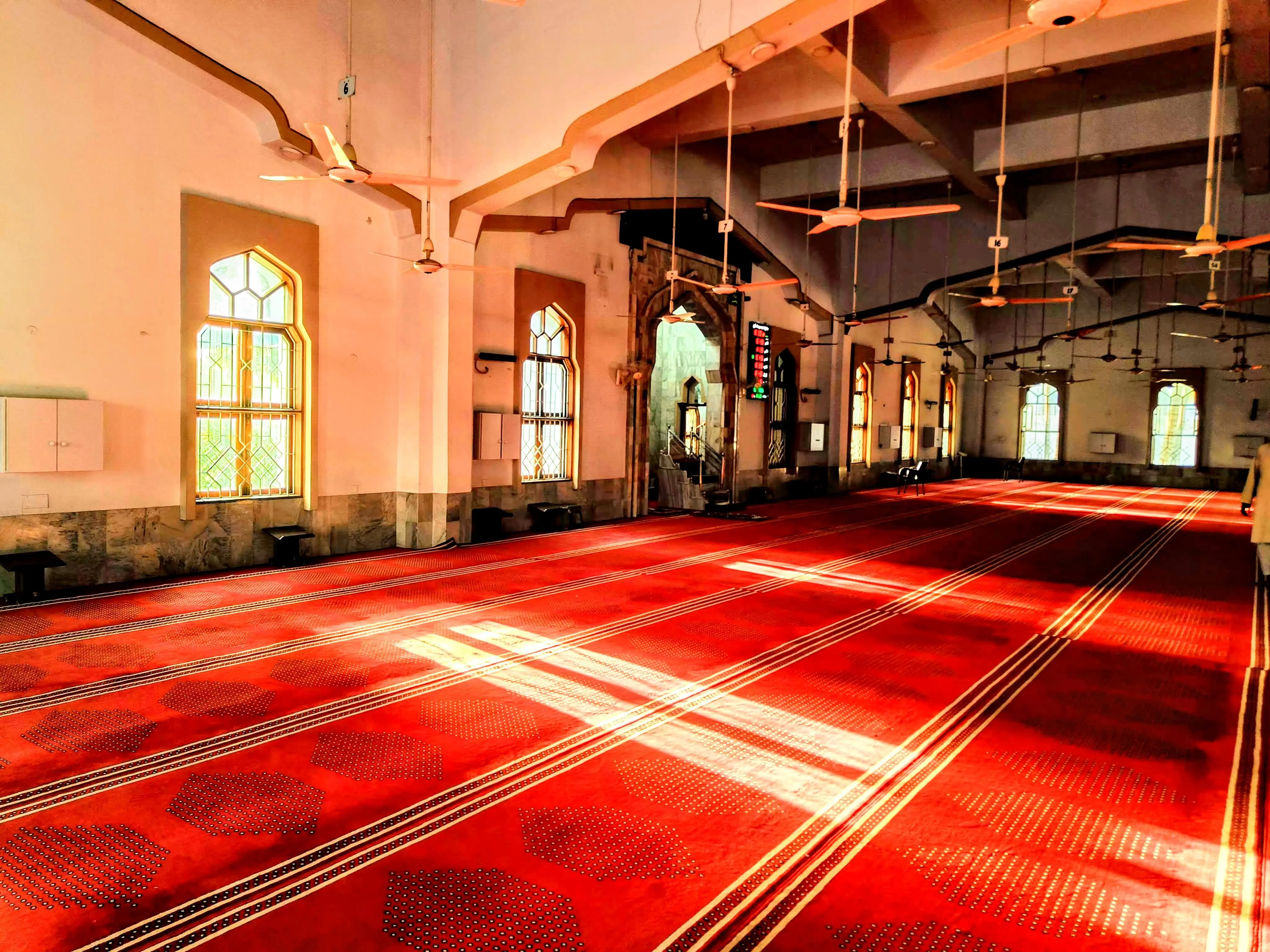 Morning light streaming through arched windows across the red carpet of Bait ul Mukarram prayer hall — mimbar visible in the distance