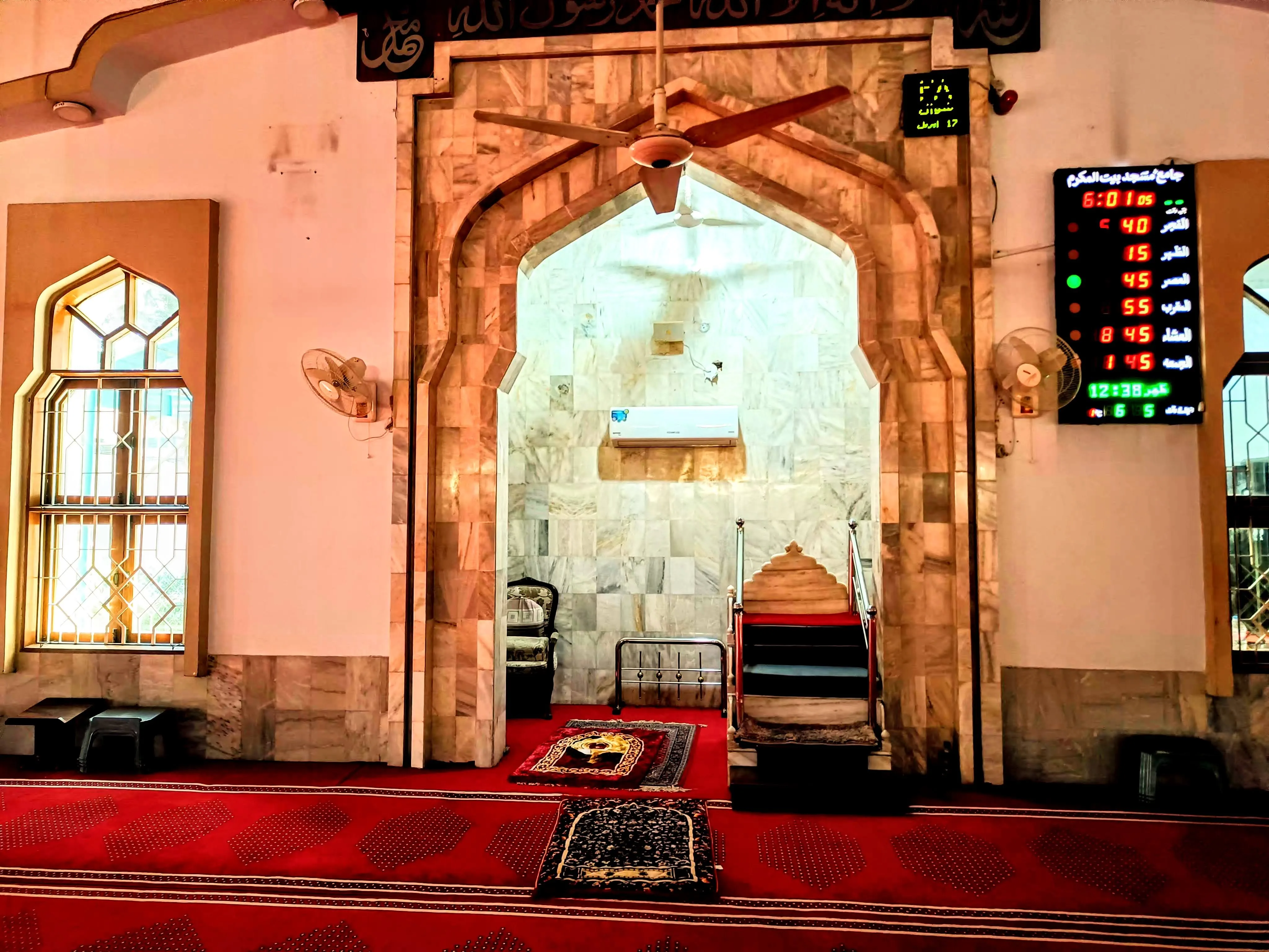 The mihrab and mimbar of Jamia Masjid Bait ul Mukarram — marble arch, mimbar steps, prayer time display board, and Kalimah inscribed above